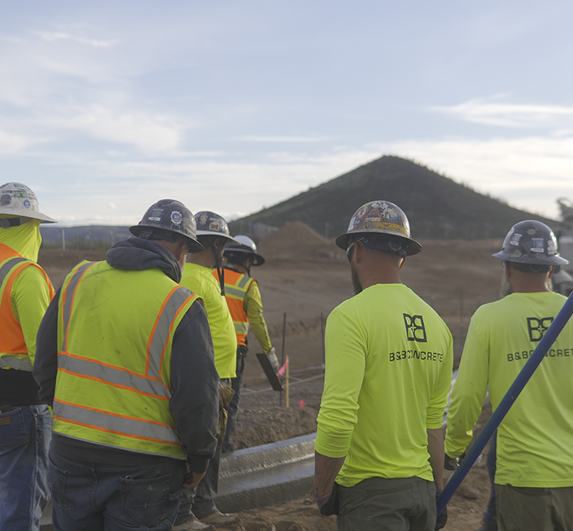A group of construction workers in safety vests and hard hats stands at a worksite with a dirt hill and pipes in the background.