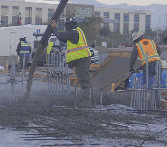 Construction workers in safety gear pour concrete onto a reinforced foundation at an outdoor building site.