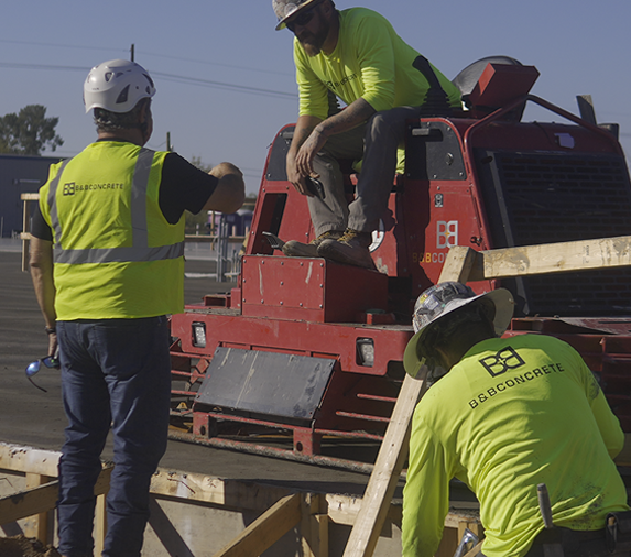 Three construction workers in safety gear and neon shirts work near a red machine at a building site, assembling wooden framework.