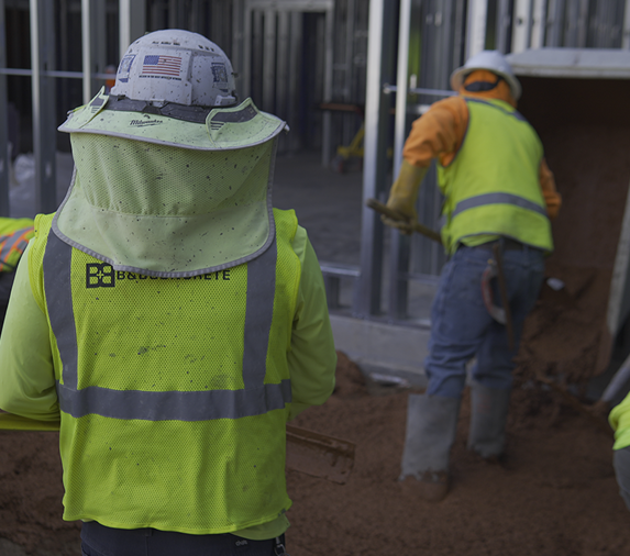 Construction workers wearing safety gear pour and spread concrete at a building site; one worker's vest displays a company logo and an American flag sticker on their helmet.