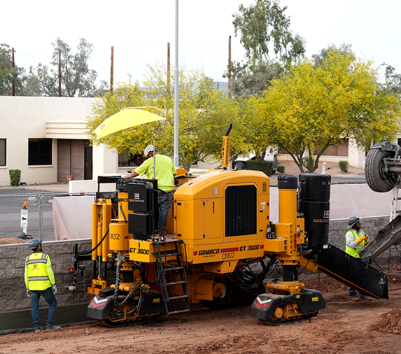 Workers operate a large yellow Gomaco GT-3600 curb machine on a construction site, with one worker using a control panel and others observing nearby.